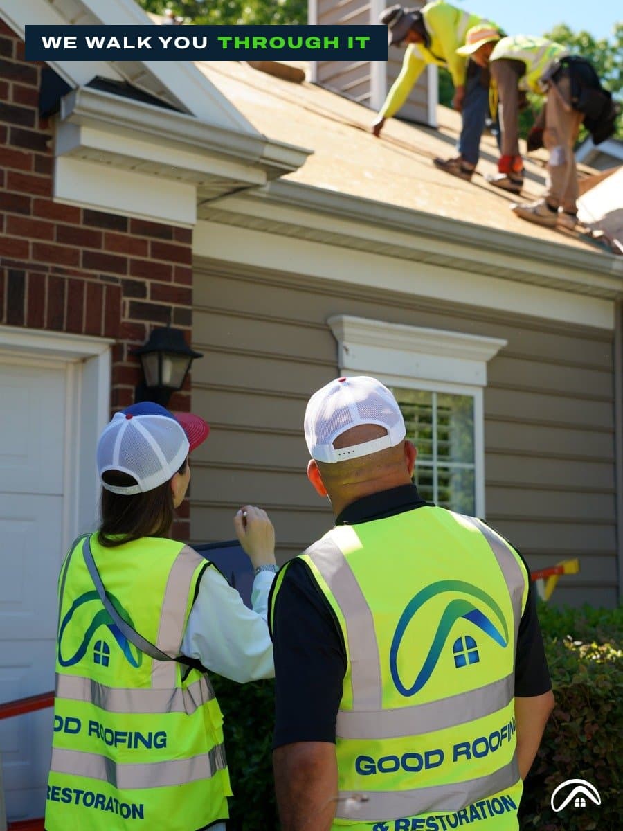 Two Good Roofing project managers in safety vests reviewing a tablet while the crew works on the roof
